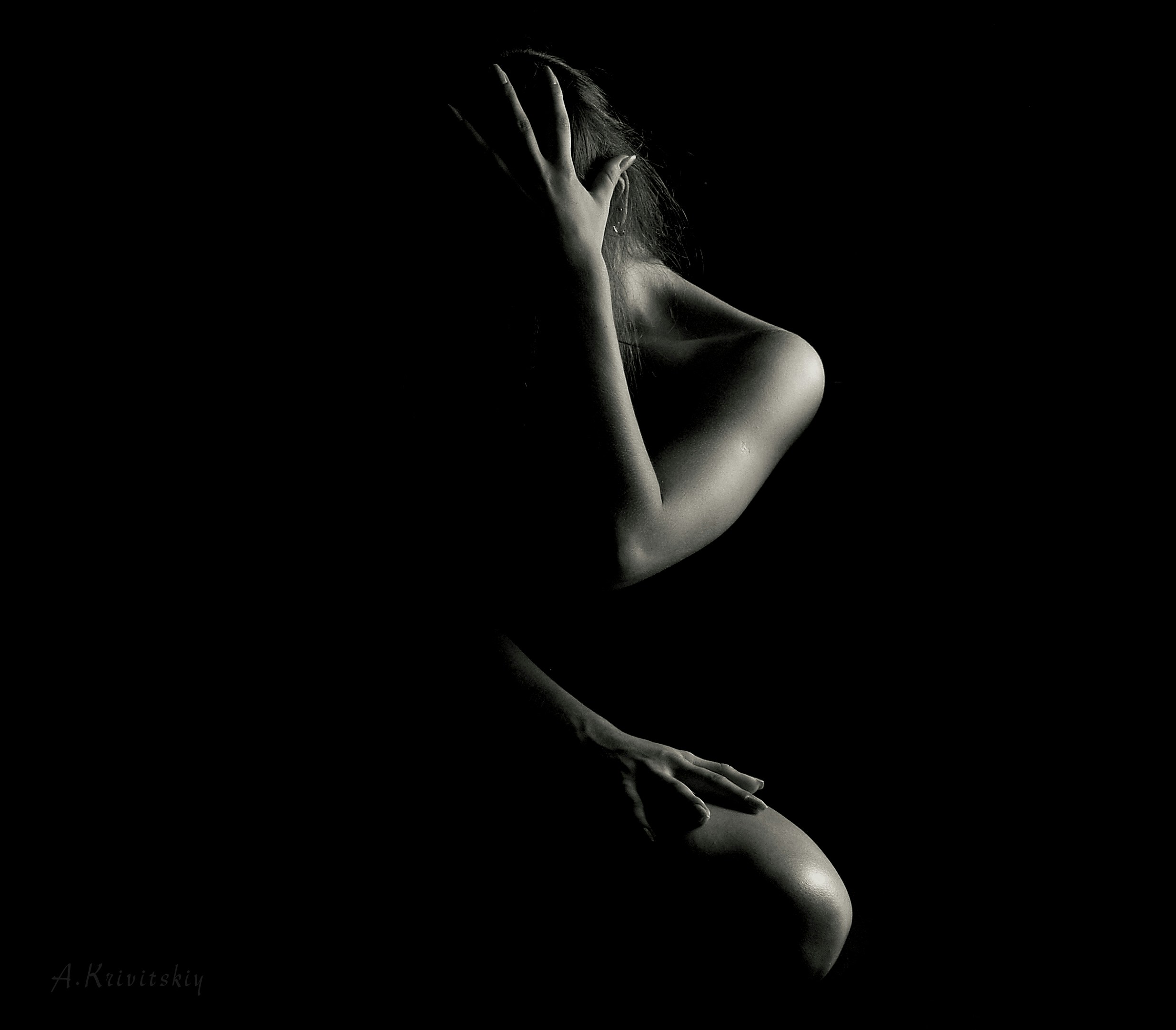 Black and white fine art photo of a nude woman in shadow, sitting with one hand covering her face, symbolizing introspection, shadow work, and emotional vulnerability
