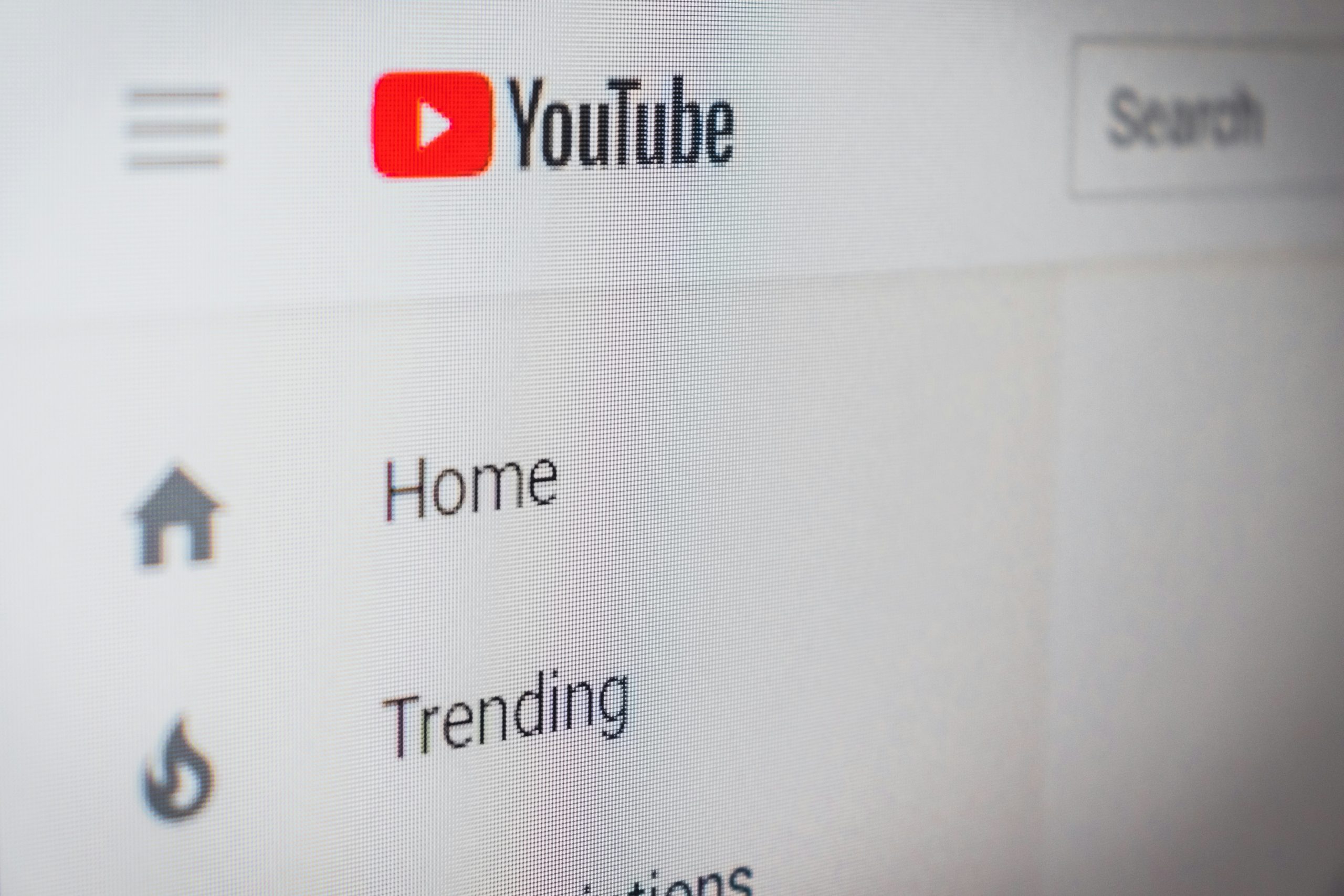 Close-up macro shot of a computer screen displaying the YouTube homepage interface, with the red and black YouTube logo in the top left, a search bar in the top right, and the sidebar menu showing Home and Trending icons; the pixel grid of the display is visible from the close angle.