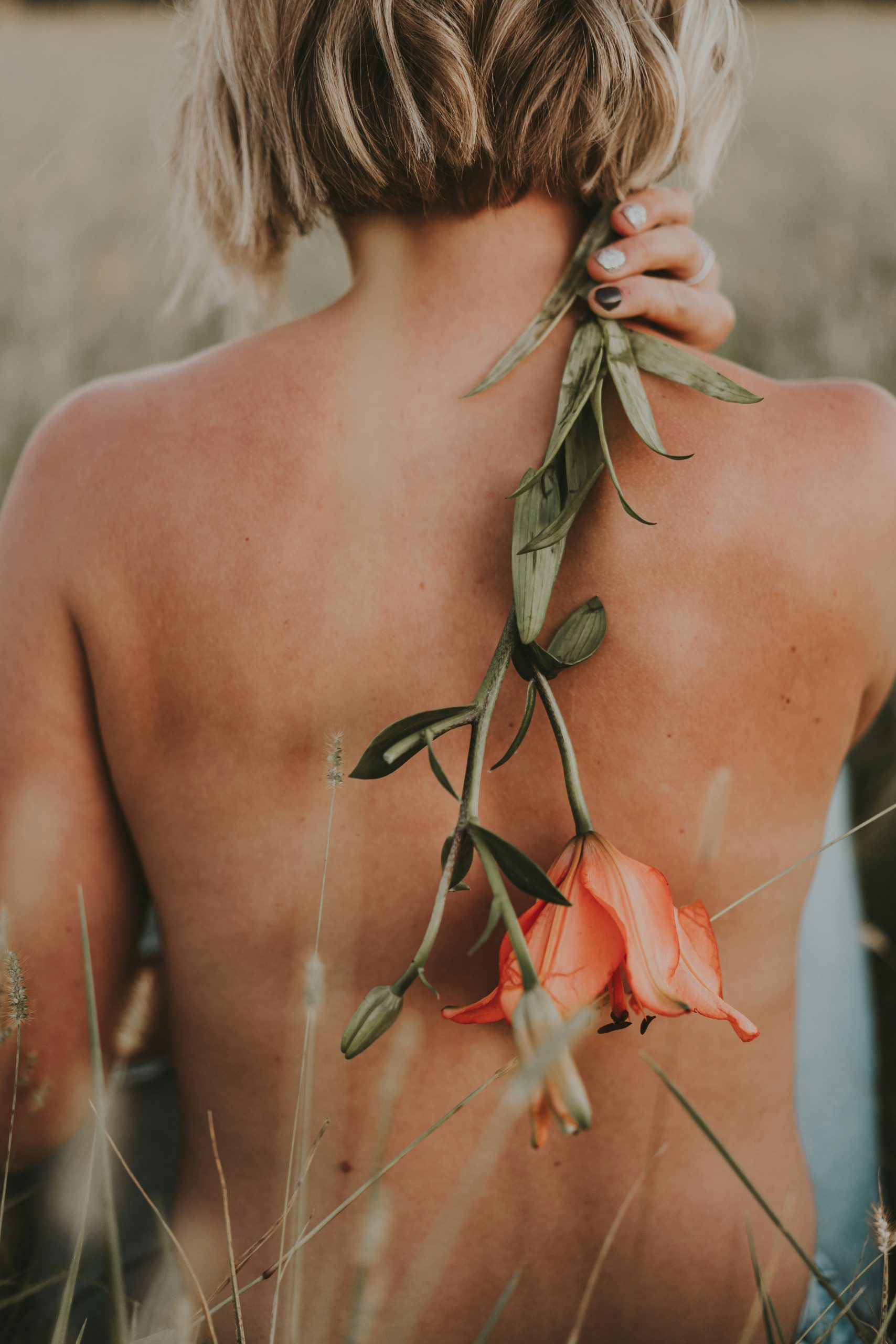 A close-up of a woman’s bare back in a natural outdoor setting, with short blonde hair. She holds a long green stem with orange flowers draped down her spine, surrounded by soft grasses, creating a calm, earthy, and organic aesthetic for somatic leadership.